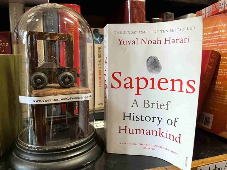 The image shows the book Sapiens by Yuval Noah Harari next to a glass dome containing a miniature chair on which rests a small wheeled trolley. Books are visible in the background.