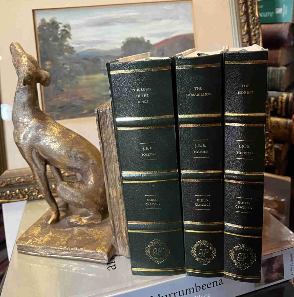 Three leather-bound books by J.R.R. Tolkien stand upright beside a bronze dog statue. A framed painting is visible in the background. 