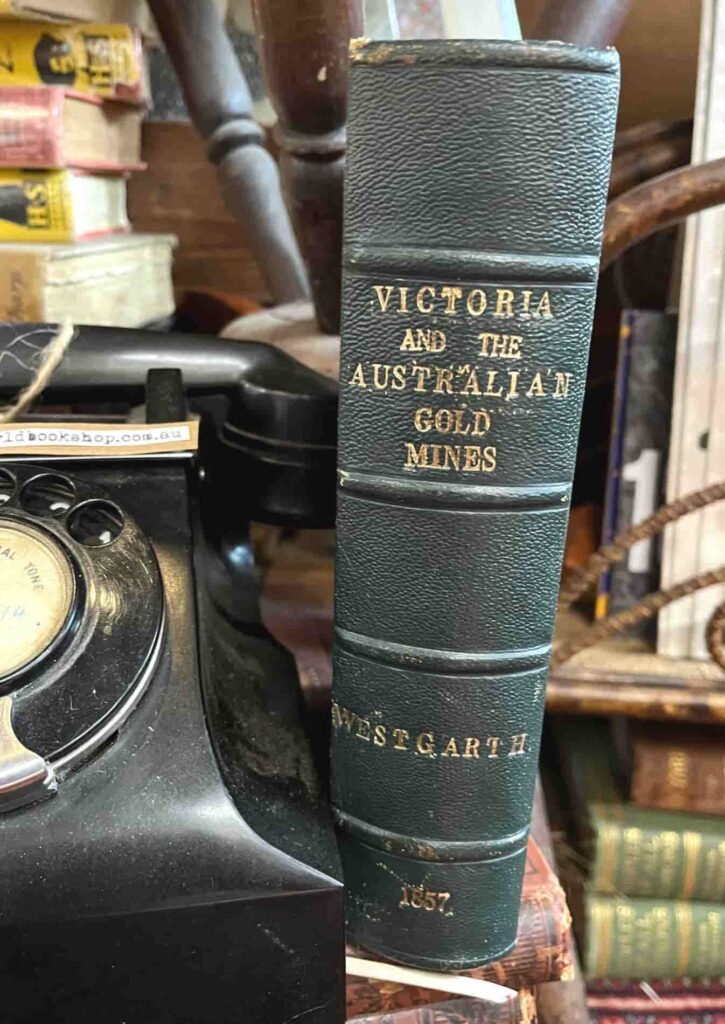 A vintage book titled "Victoria and the Australian Gold Mines" sits beside a black rotary phone. The book's spine reads Westgarth, 1857 The background features stacked old books, conveying a nostalgic, historical ambiance.