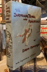 The book The Surprising Travels & Adventures of Baron Munchausen stands atop 3 other large secondhand books